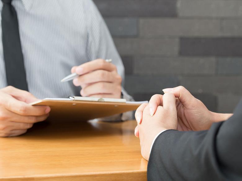 A close-up of two people in a professional setting, with one holding a clipboard and pen, while the other has hands clasped, likely during an interview or meeting.